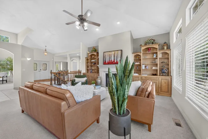 Vaulted ceilings with a ceiling fan, a fireplace, wood-finish built-in shelving, large windows with blinds, and a wood staircase with a white baluster