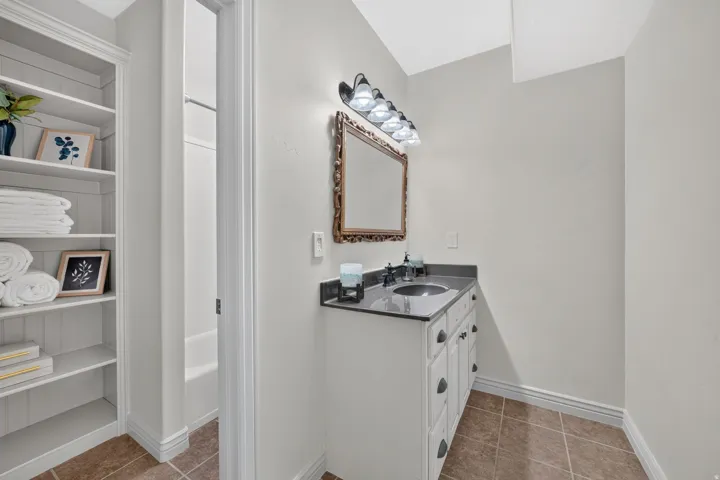 Built-in shelving, white vanity with dark countertop, undermount sink, overhead vanity lighting, framed wall mirror, and tile flooring