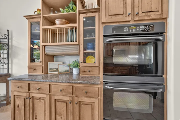 Custom wood cabinetry with glass-front display cabinets, stone countertops, and built-in double ovens