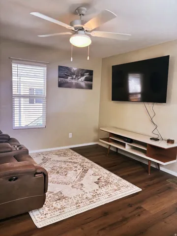 Wood-finish flooring, ceiling fan with integrated lighting, window with blinds, wall-mounted display, and light-toned wall paint