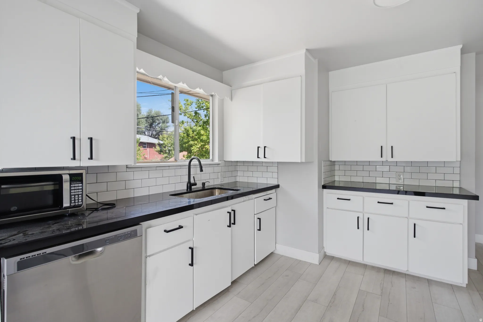 White cabinetry with black hardware, dark countertops, a subway tile backsplash, and light wood-finish flooring