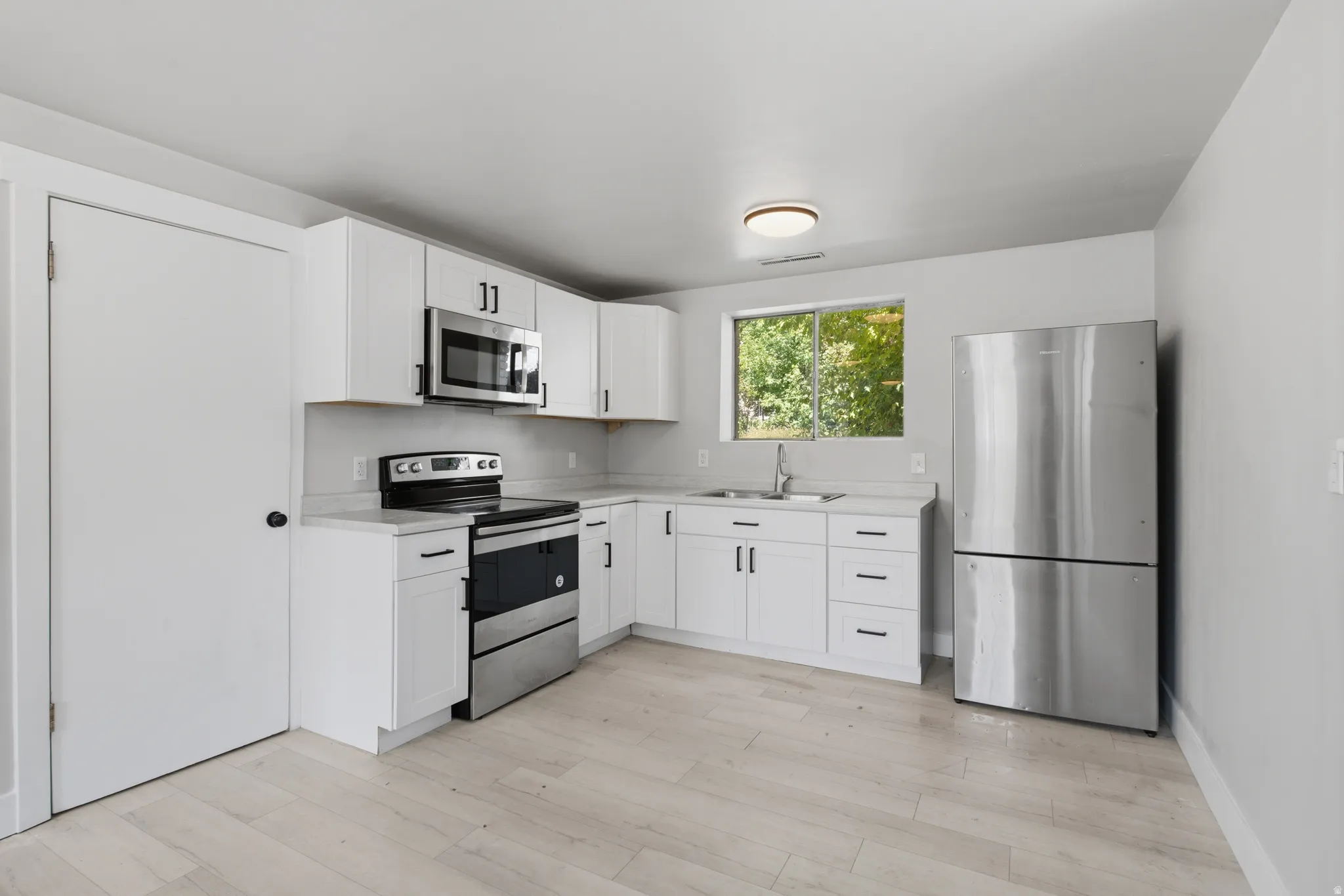 White cabinetry with matte black hardware, stainless steel appliances, light-toned flooring, and a window
