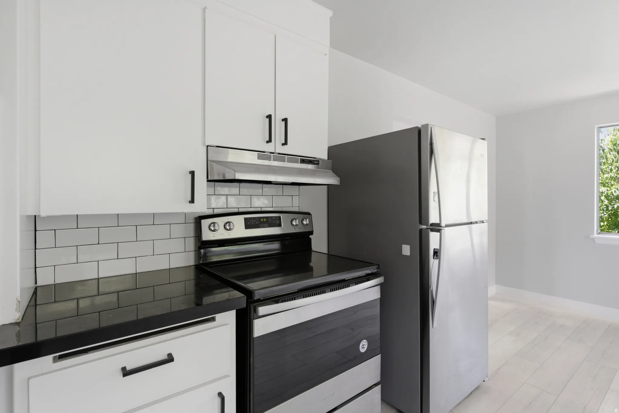 White cabinetry with matte black hardware, dark countertops, subway tile backsplash, stainless steel appliances, and light wood-finish flooring