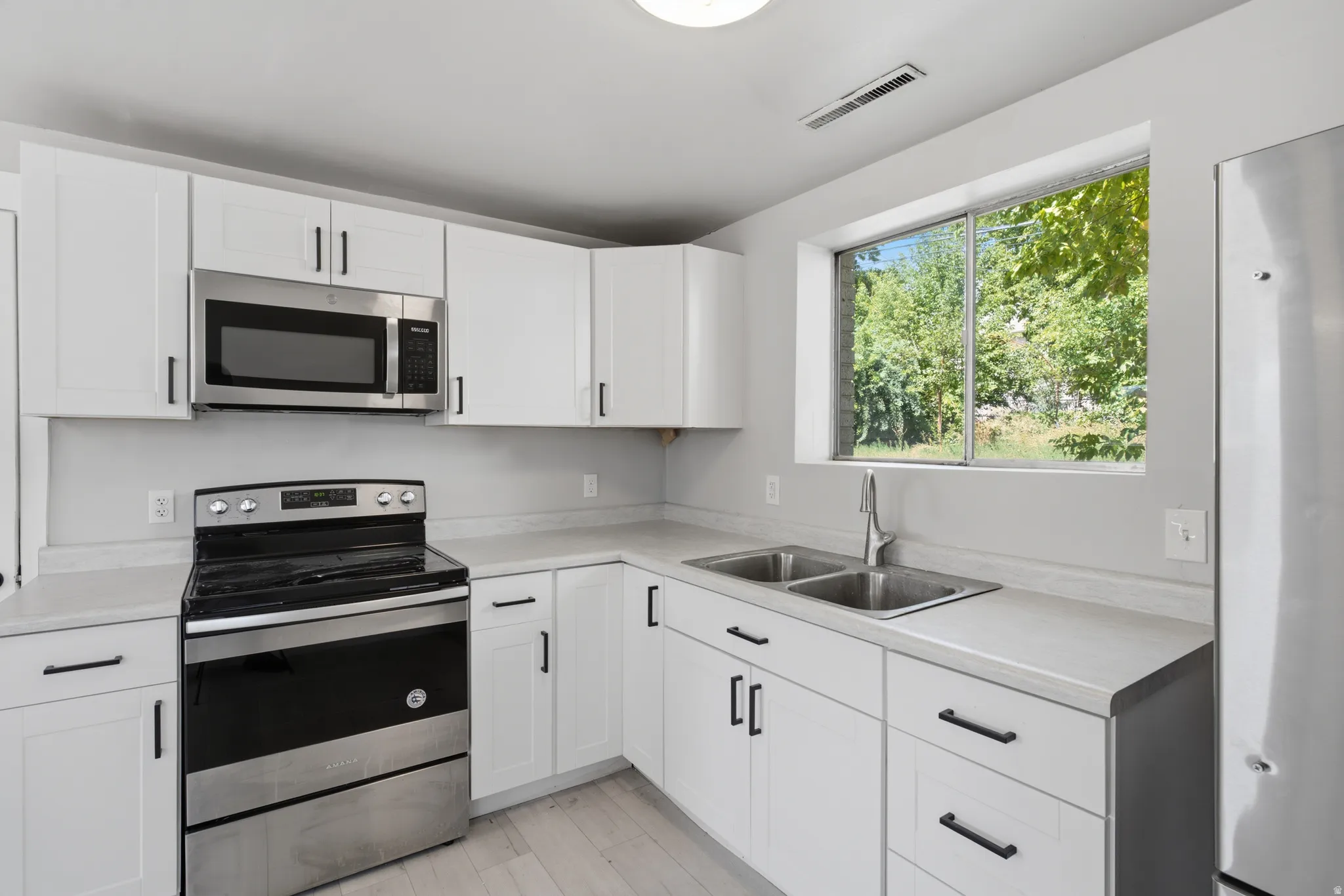 White shaker-style cabinetry with black hardware, light-toned countertops, stainless steel appliances, and a dual-basin sink with a gooseneck faucet