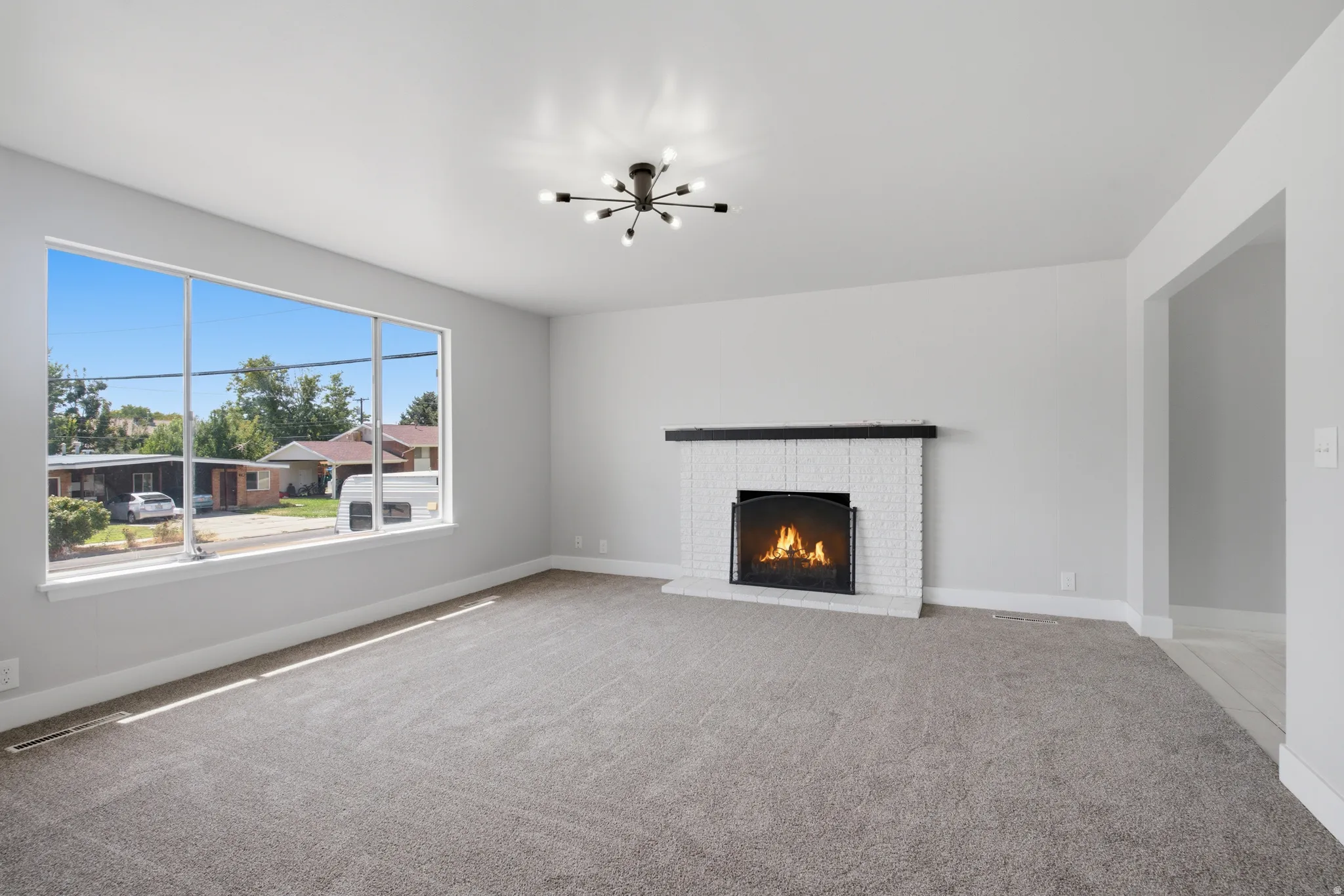 Spacious living area featuring a large picture window, a white brick fireplace with a dark wood mantel, and a contemporary ceiling light fixture