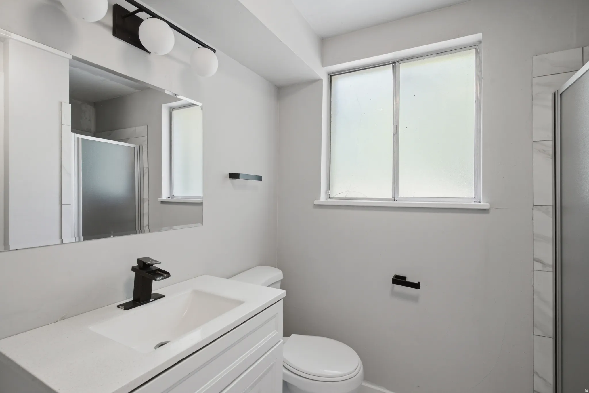 Vanity with integrated sink and matte black faucet, wall-mounted light fixture, and tiled shower enclosure