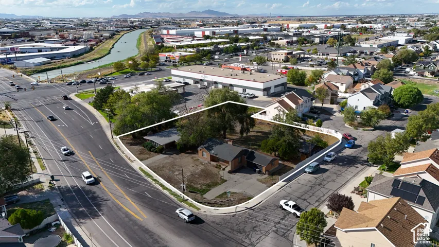 Aerial perspective of corner lot featuring single-level structure, multi-pitch roofline, mature trees, and paved driveway