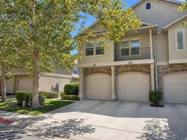 Elegant townhome featuring one-car garage with arched brick detailing, neutral stucco and stone exterior, mature shade trees providing natural landscaping, manicured shrub plantings, and wide concrete driveway