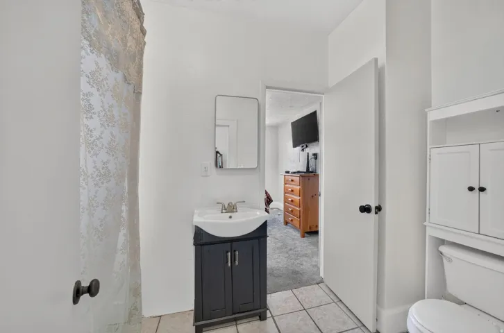 Tile flooring, single vanity with dark cabinetry, wall-mounted mirror, white storage cabinet, and toilet