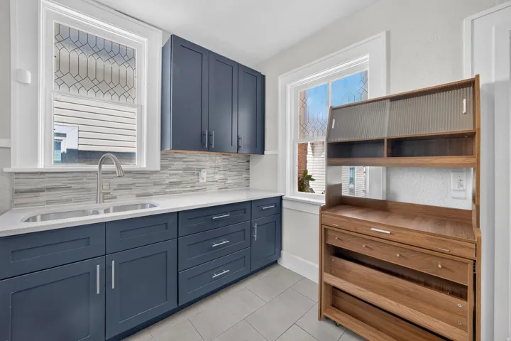 Navy cabinetry, white countertops, stainless steel sink, mosaic tile backsplash, and light gray tile flooring