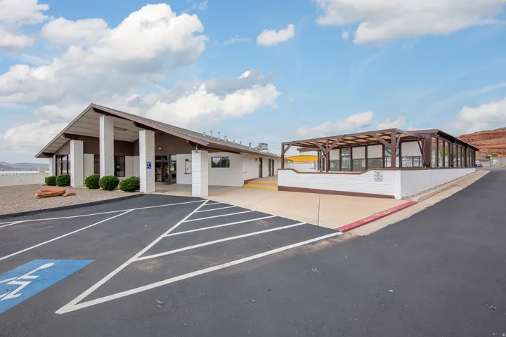 Paved parking lot with designated accessible parking, covered building entrance with white columns, low-maintenance landscaping, and an enclosed outdoor patio area with a wooden pergola
