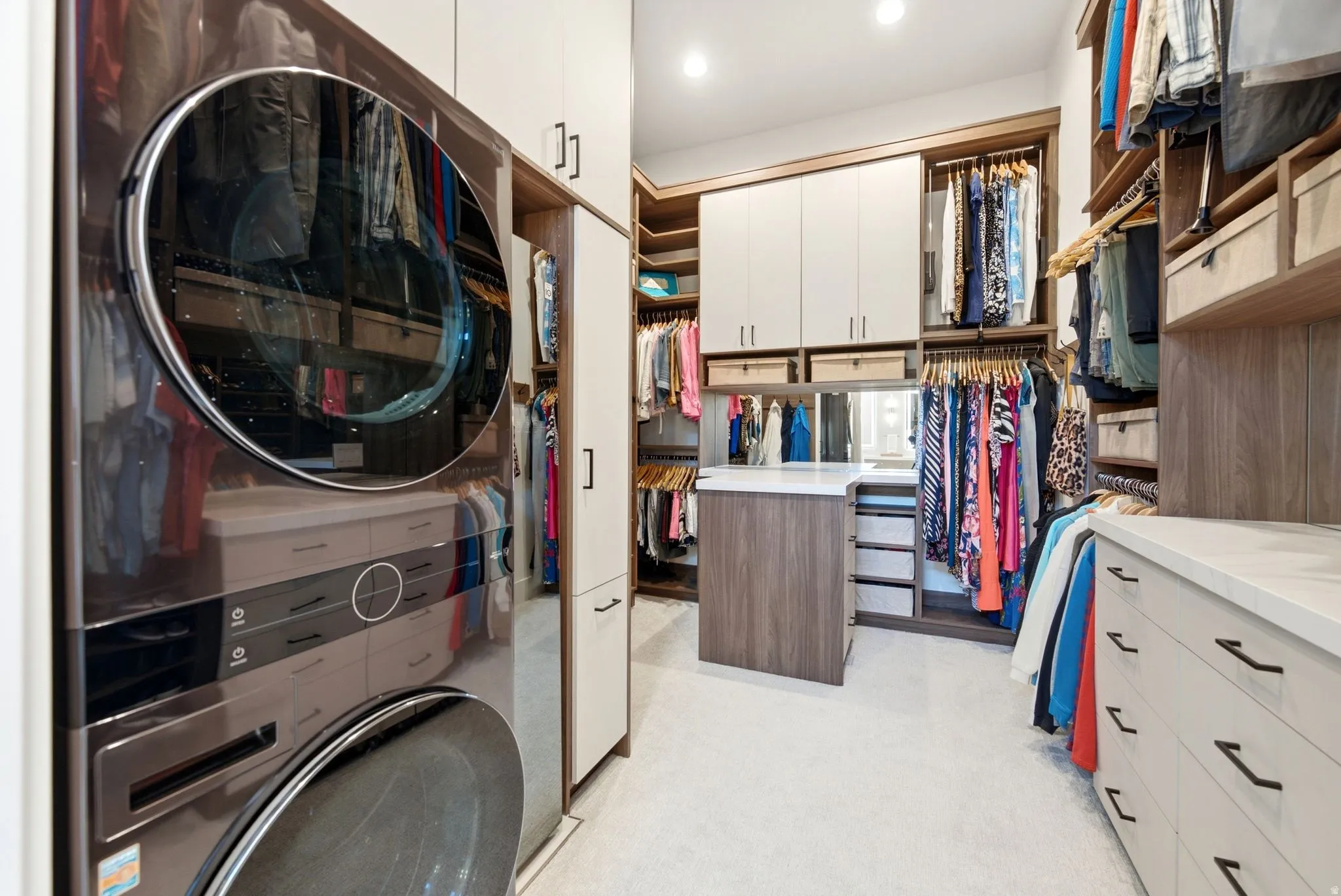 Luxurious walk-in closet featuring custom cabinetry with white upper storage and wood-finish lower drawers, a central island with built-in shelving for organized wardrobe display, abundant natural lighting, and a stacked washer-dryer unit integrated into