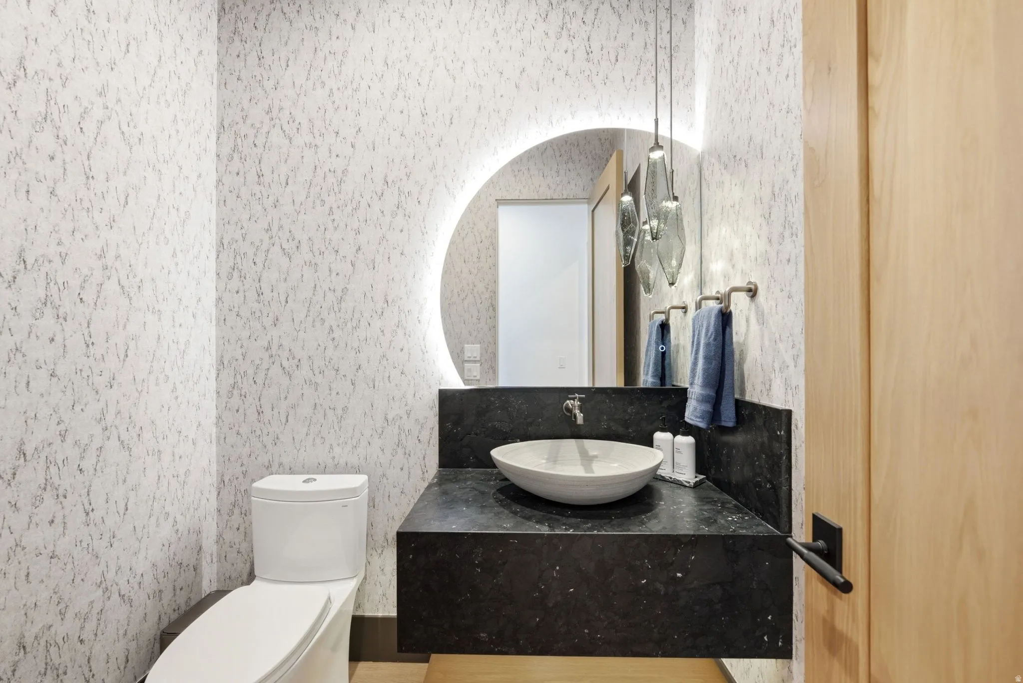 Floating vanity with a dark countertop and vessel sink, illuminated circular mirror, and textured wall covering