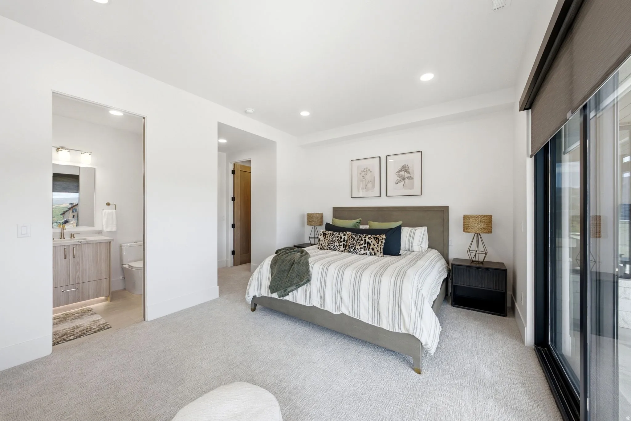 Recessed lighting, light-toned carpeting, a sliding glass door with a privacy shade, and an en-suite bathroom with a wood-finish vanity and integrated sink