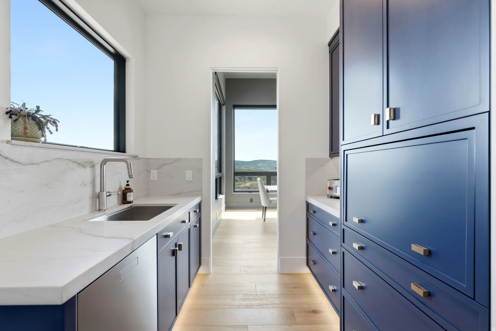 Galley kitchen featuring light wood-finish flooring, deep blue cabinetry with brass hardware, a stainless steel sink, and light-toned countertops with a matching backsplash