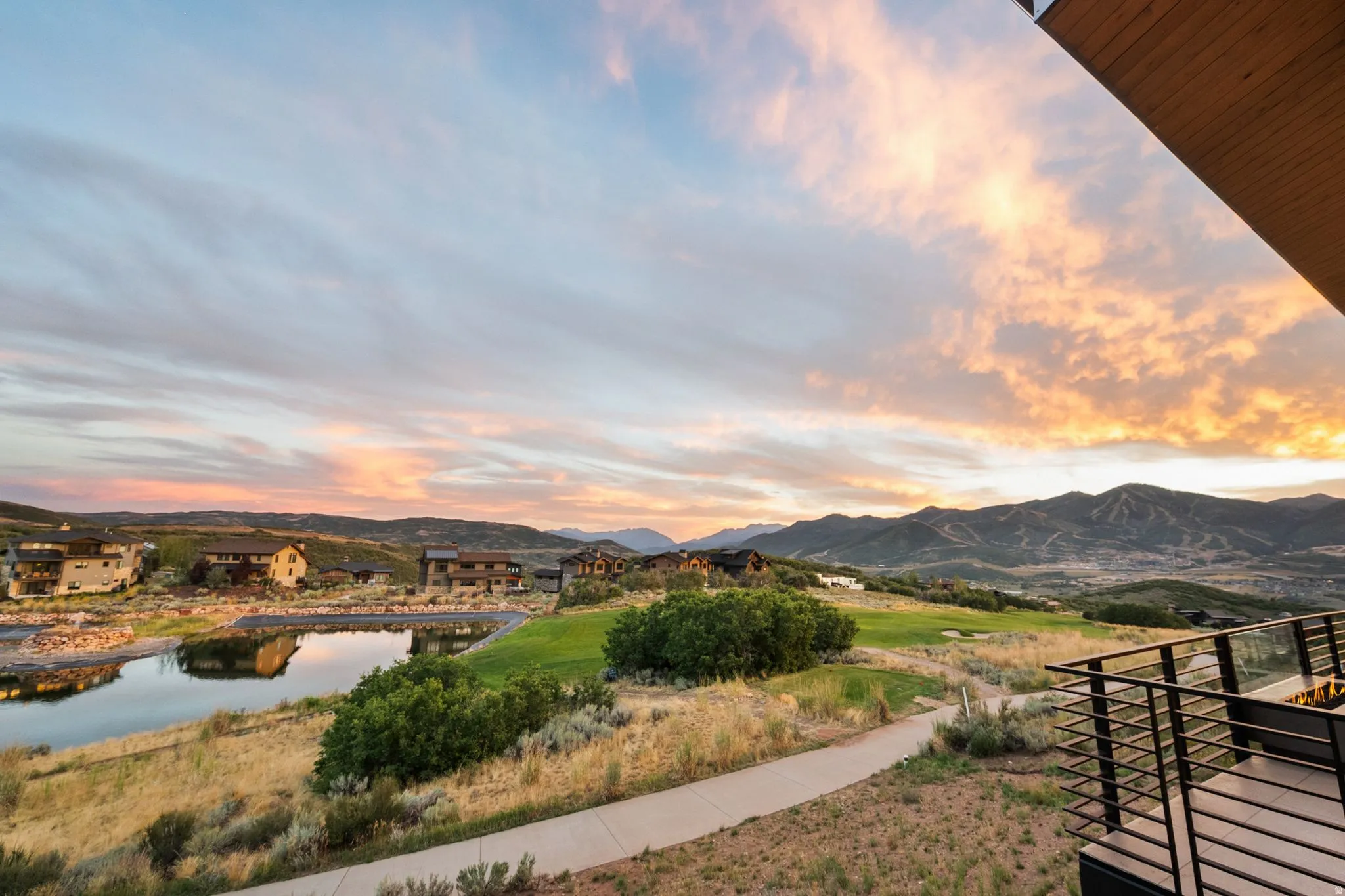 Expansive outdoor balcony with glass and metal railing, overlooking a scenic mountain landscape, a golf course, and a reflective pond