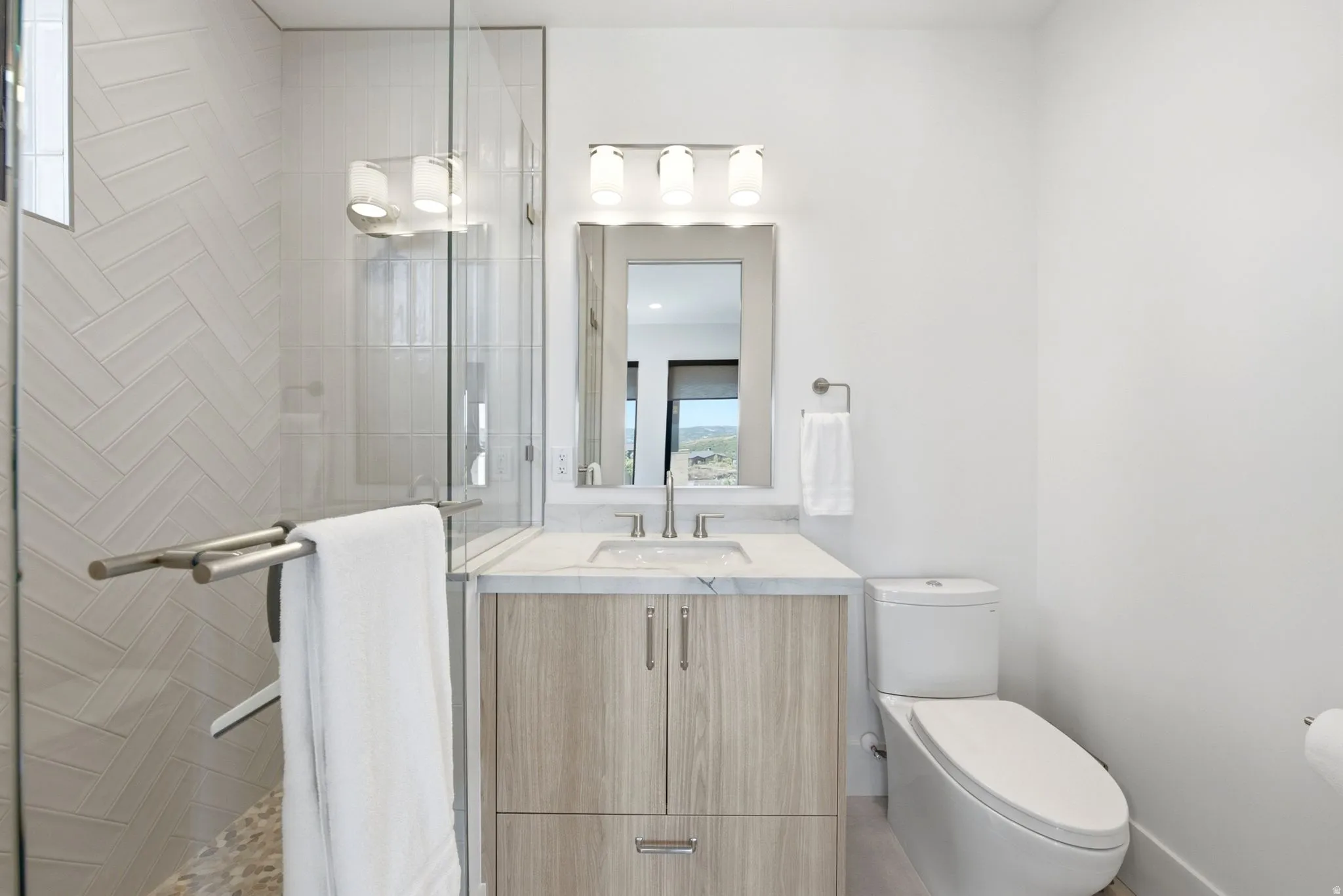 Glass-enclosed shower with herringbone tile, single vanity with light wood-finish cabinetry, white countertop, and a contemporary mirror