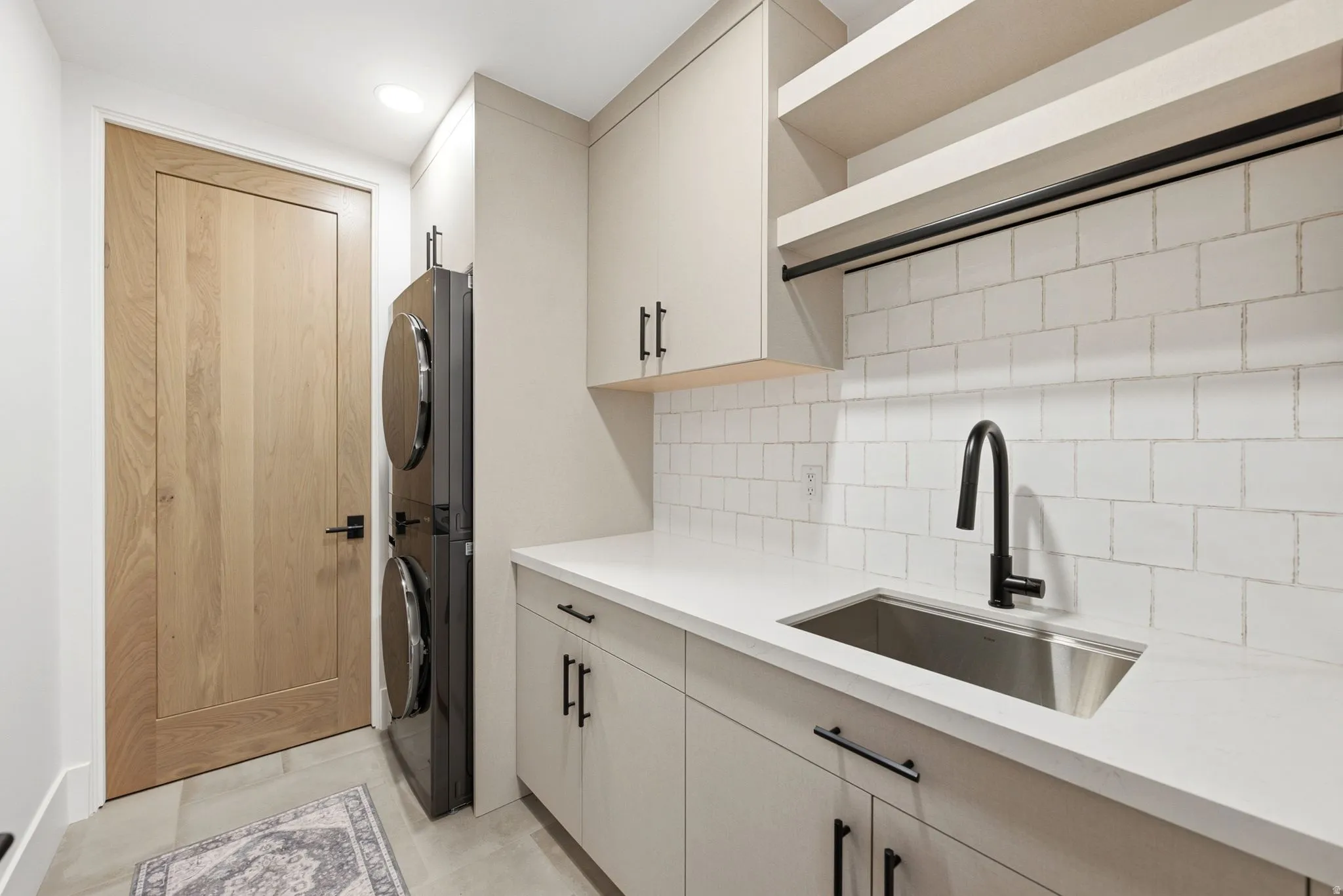 Light-toned wood door, light gray cabinetry with black hardware, white countertop, stainless steel sink with a black faucet