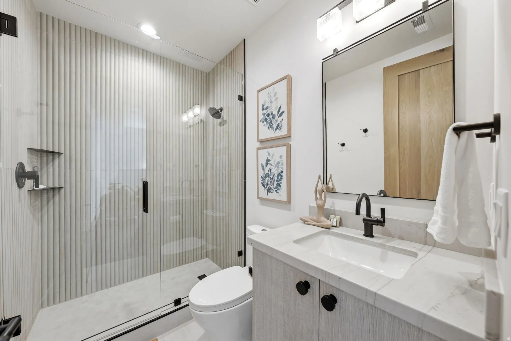 Frameless glass shower enclosure, fluted shower wall paneling, vanity with integrated sink and stone countertop, matte black fixtures, and recessed lighting