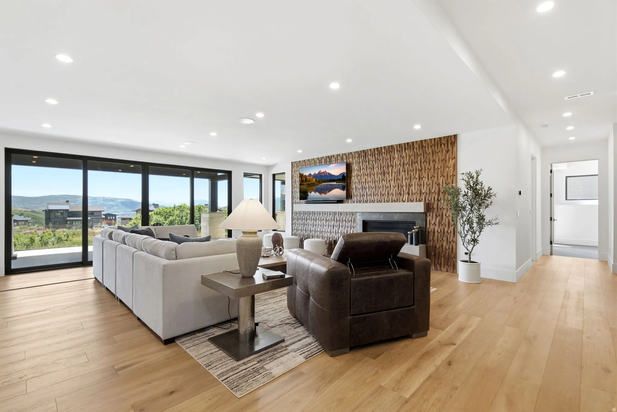 Spacious living area featuring expansive glass sliding doors, wood-finish flooring, recessed lighting, and a modern fireplace with a textured wood-panel surround