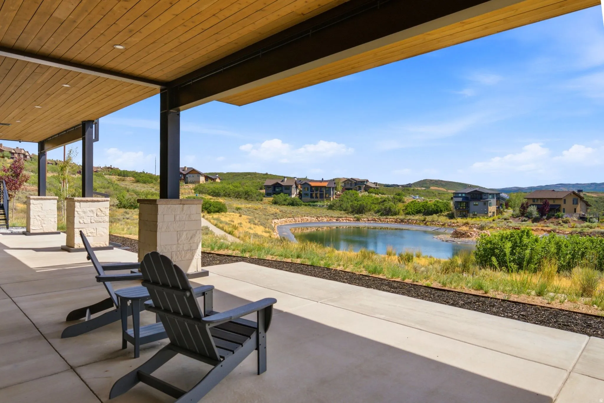 Covered patio with a wood-finish ceiling, exposed structural beams, and stone-clad pillars, overlooking a water feature and surrounding landscape