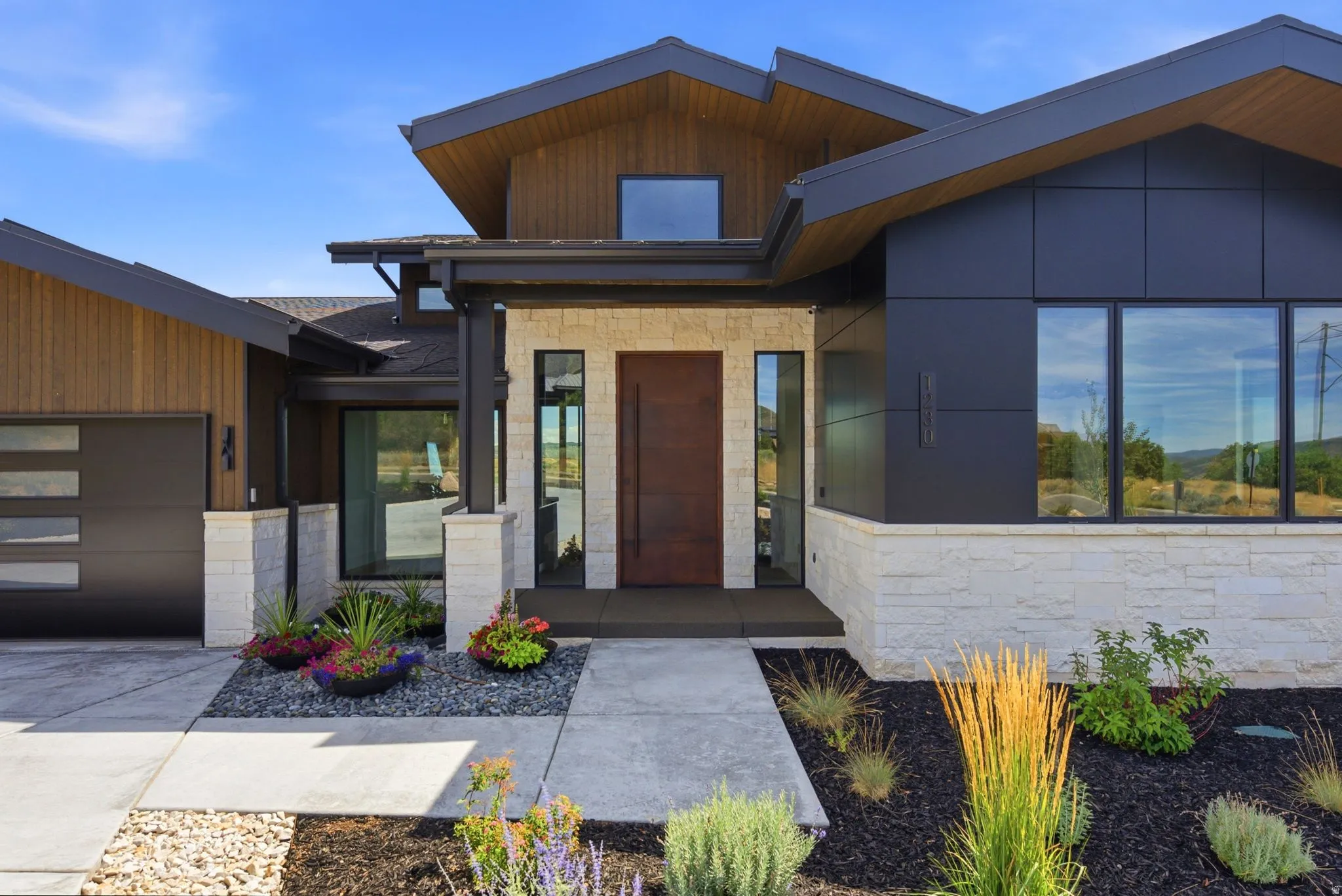 Modern exterior featuring a prominent wood entry door, stone facade accents, dark panel siding, large windows, and a segmented garage door