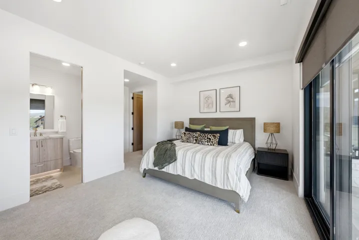 Recessed lighting, light-toned carpeting, a sliding glass door with a privacy shade, and an en-suite bathroom with a wood-finish vanity and integrated sink