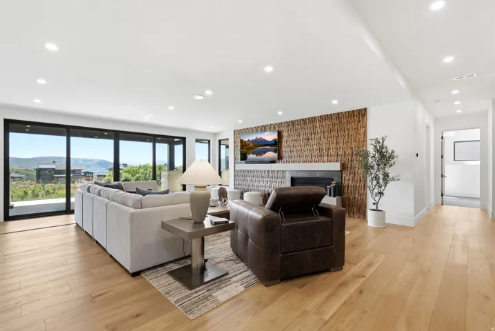 Spacious living area featuring expansive glass sliding doors, wood-finish flooring, recessed lighting, and a modern fireplace with a textured wood-panel surround