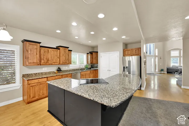Chef's kitchen featuring custom wood cabinetry, granite countertops, a large center island with black base and stainless steel appliances, recessed lighting throughout, and hardwood flooring that extends into the adjoining living spaces
