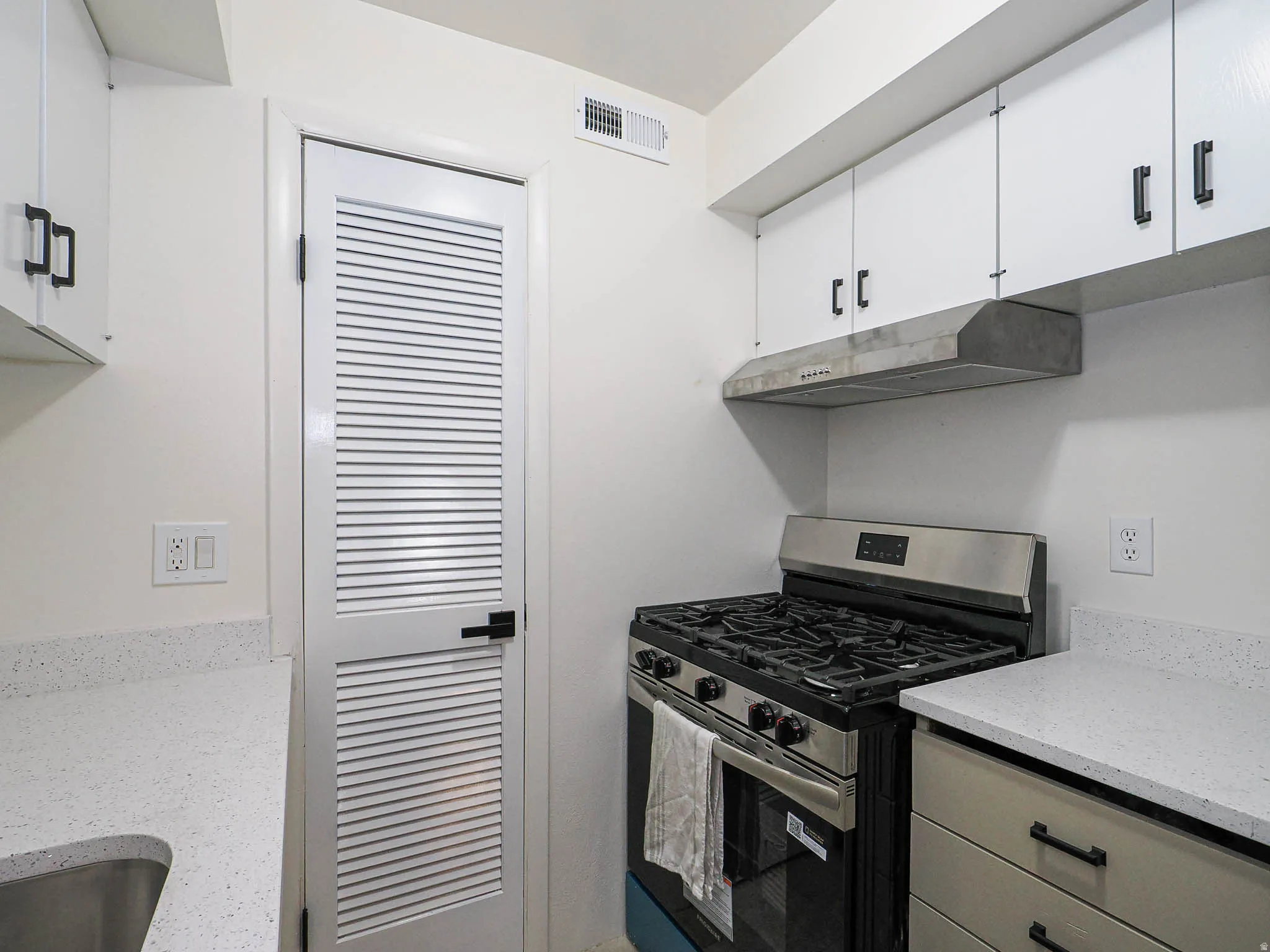 White cabinetry with matte black hardware, light-toned countertops, stainless steel range with hood, and a louvered door