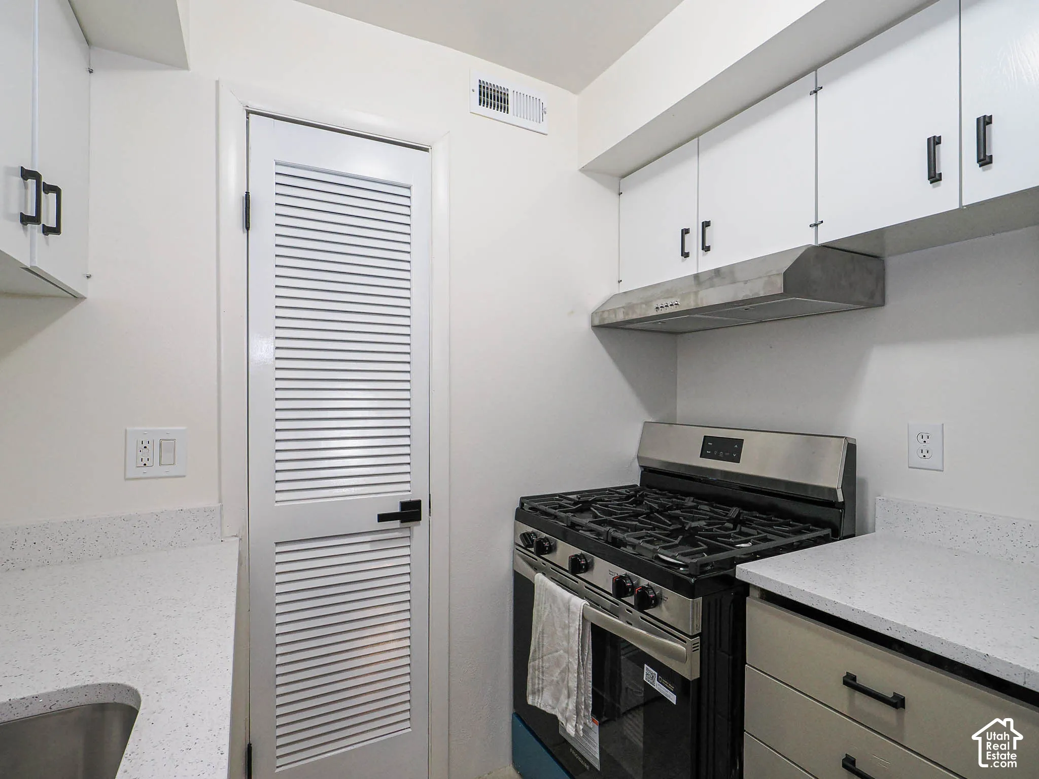 White cabinetry with matte black hardware, light-toned countertops, stainless steel range with hood, and a louvered door