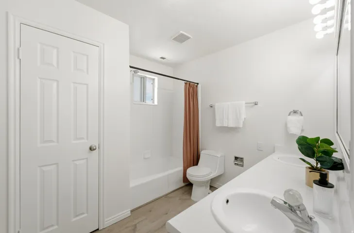 Dual vanity with integrated sinks, modern wall-mounted light fixture, white six-panel door, wood-finish flooring, and a window within the shower enclosure