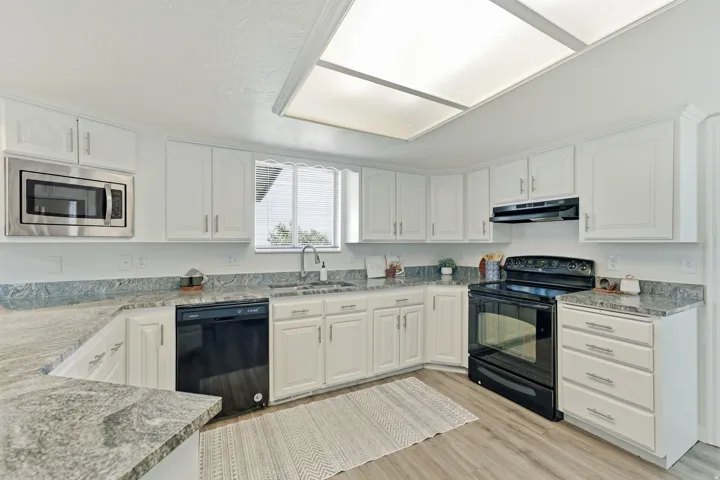 White cabinetry, light-toned wood-finish flooring, stainless steel microwave, black range, and gray flecked countertops