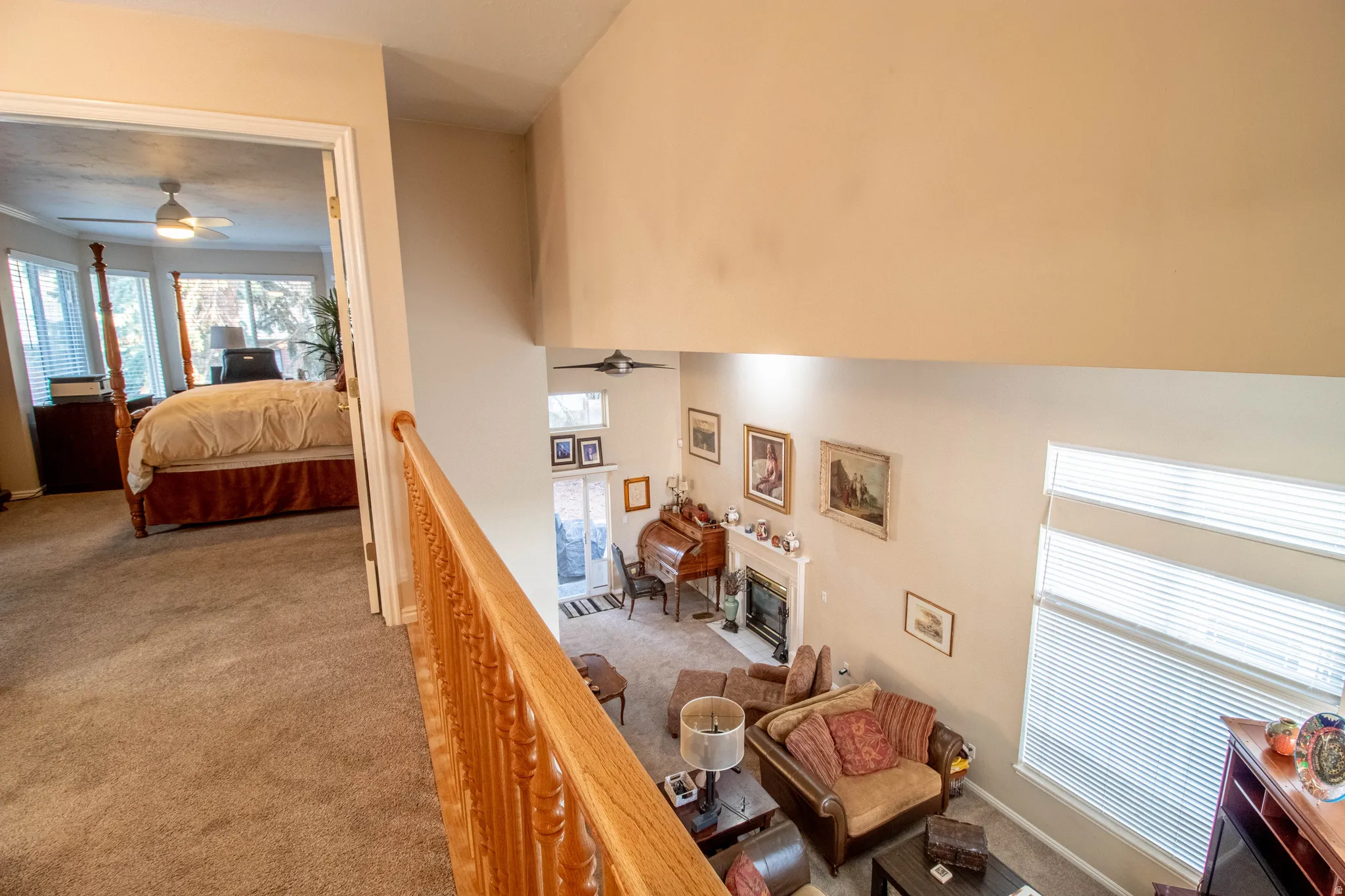 Carpeted hallway with wood banister overlooking a living space with a fireplace, high ceilings, and large windows