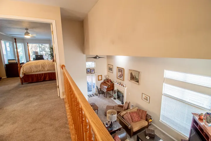 Carpeted hallway with wood banister overlooking a living space with a fireplace, high ceilings, and large windows