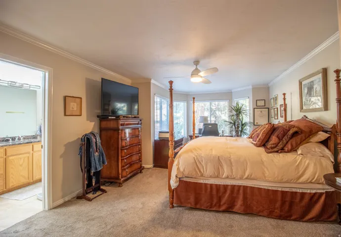Carpeted bedroom featuring a ceiling fan with integrated lighting, crown molding, and a bay window