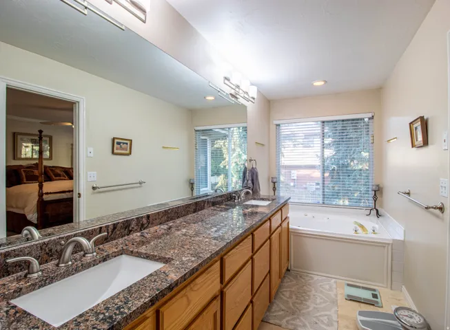 Dual vanity with integrated sinks, expansive counter space with a stone-finish surface, wood-finish cabinetry, and a built-in jetted tub