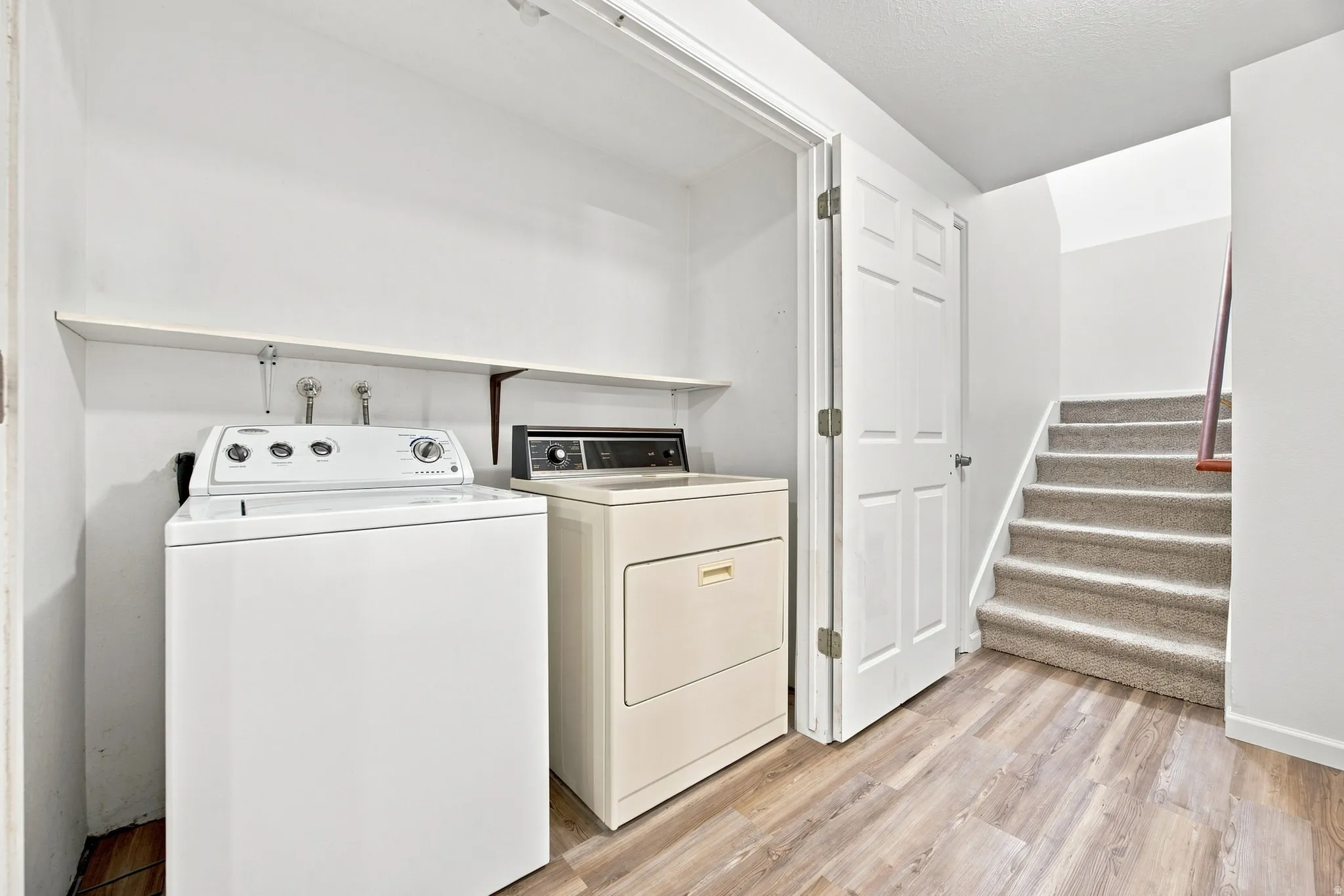 Dedicated laundry area featuring a wall shelf, wood-finish flooring, and a carpeted staircase