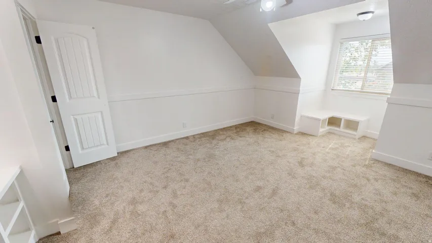 Window with blinds, wall-mounted light fixture, angled wall design, built-in shelving, and carpet flooring