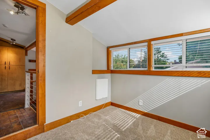 Carpeted flooring, wood-framed windows with blinds, exposed wood beams, and a wall-mounted heating unit