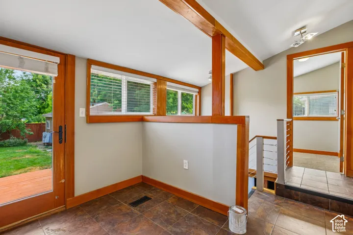 Terracotta-toned tile flooring, wood-framed glass door, multiple windows with blinds, exposed wood beams, and a stairwell with contemporary metal railing