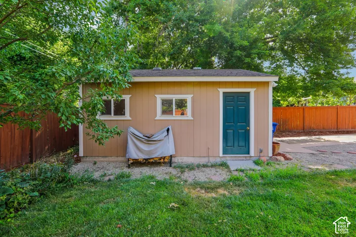 Detached structure featuring tan siding, a dark teal door, two windows with white trim, and a shingle roof