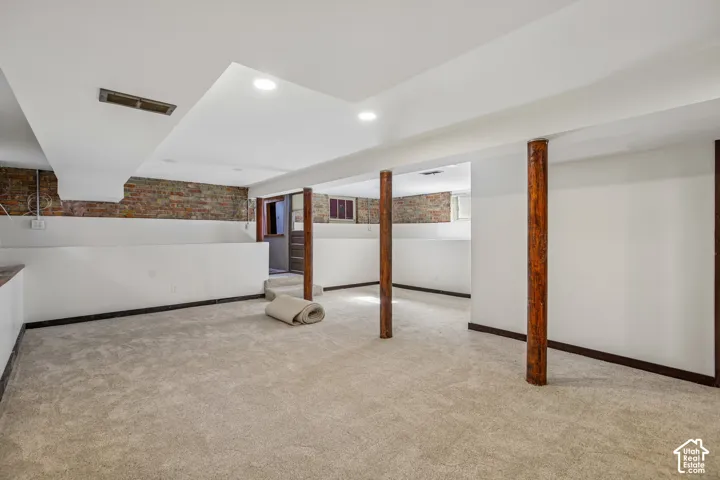 Lower level space with carpet flooring, exposed brick wall, and wood-wrapped support columns
