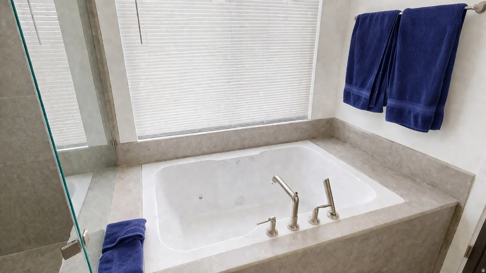 Built-in soaking tub with brushed nickel fixtures, surrounded by neutral-toned tile, and featuring a window with privacy blinds