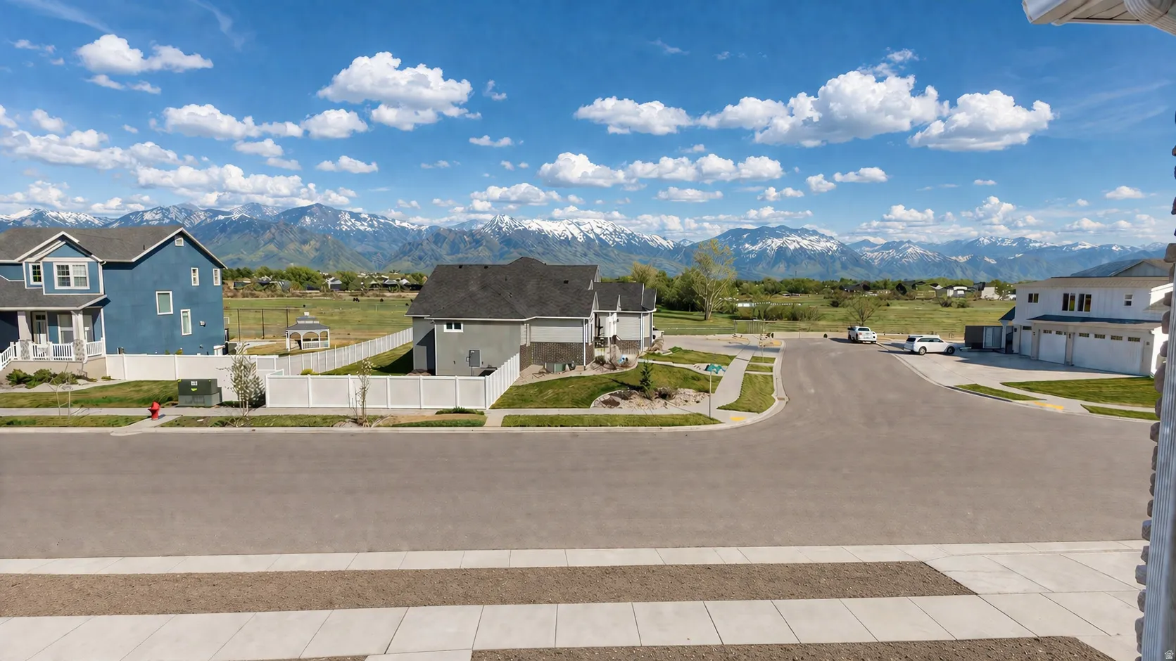 This is an Ai enhanced Photo. Snow-capped mountain range backdrop, expansive green field, neighborhood street with sidewalks, white fencing, and varied residential architecture
