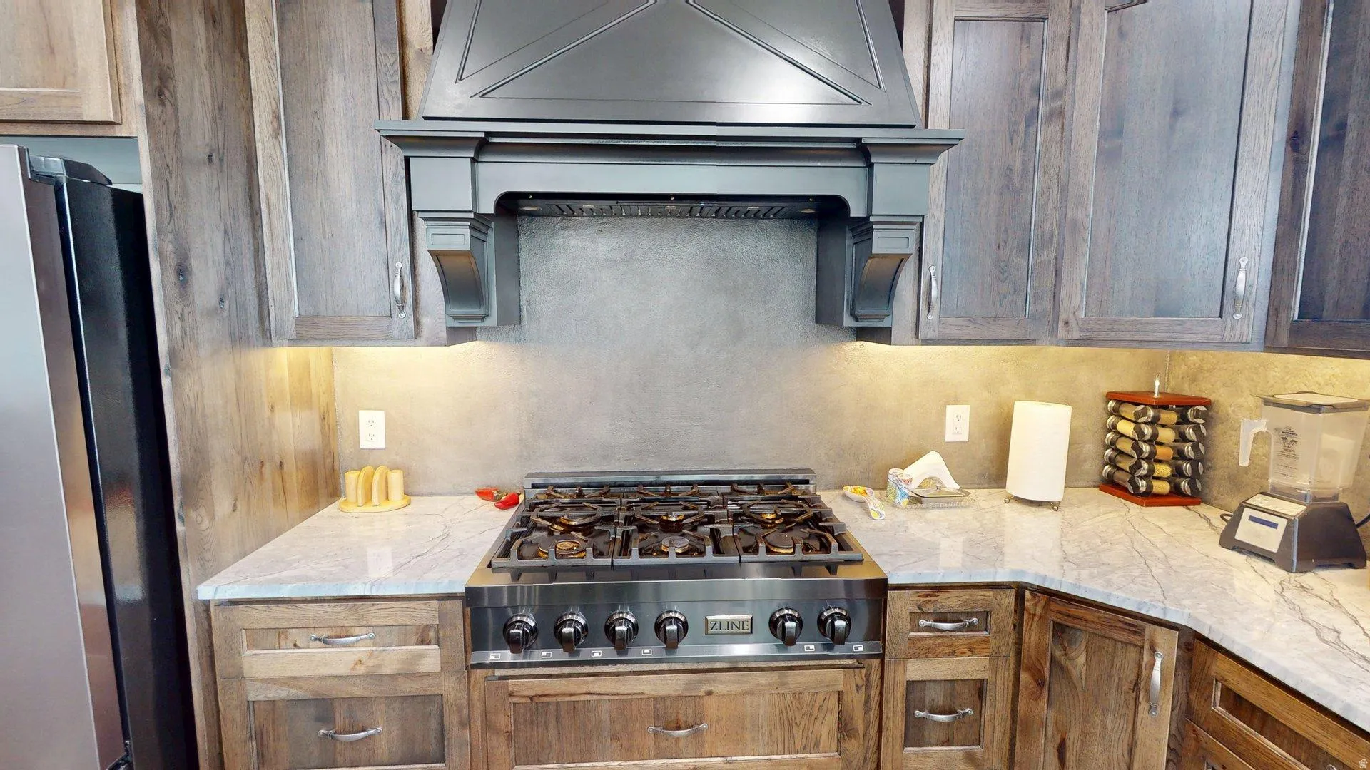 Stainless steel gas range, custom range hood, wood-finish cabinetry, light-toned stone countertops, and a full-height backsplash