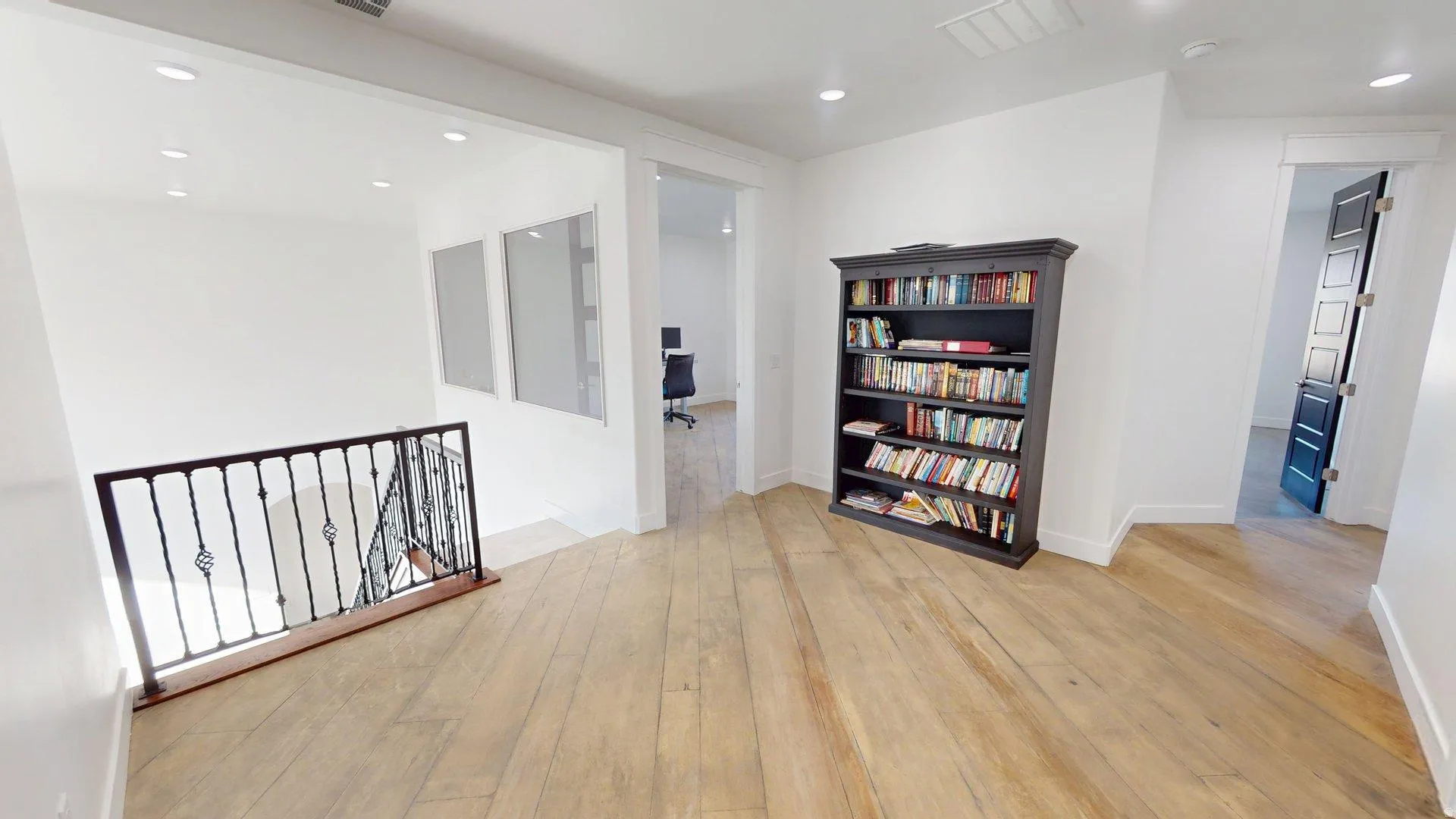 Hardwood flooring, recessed lighting, white walls, dark iron railing, and a dark paneled door