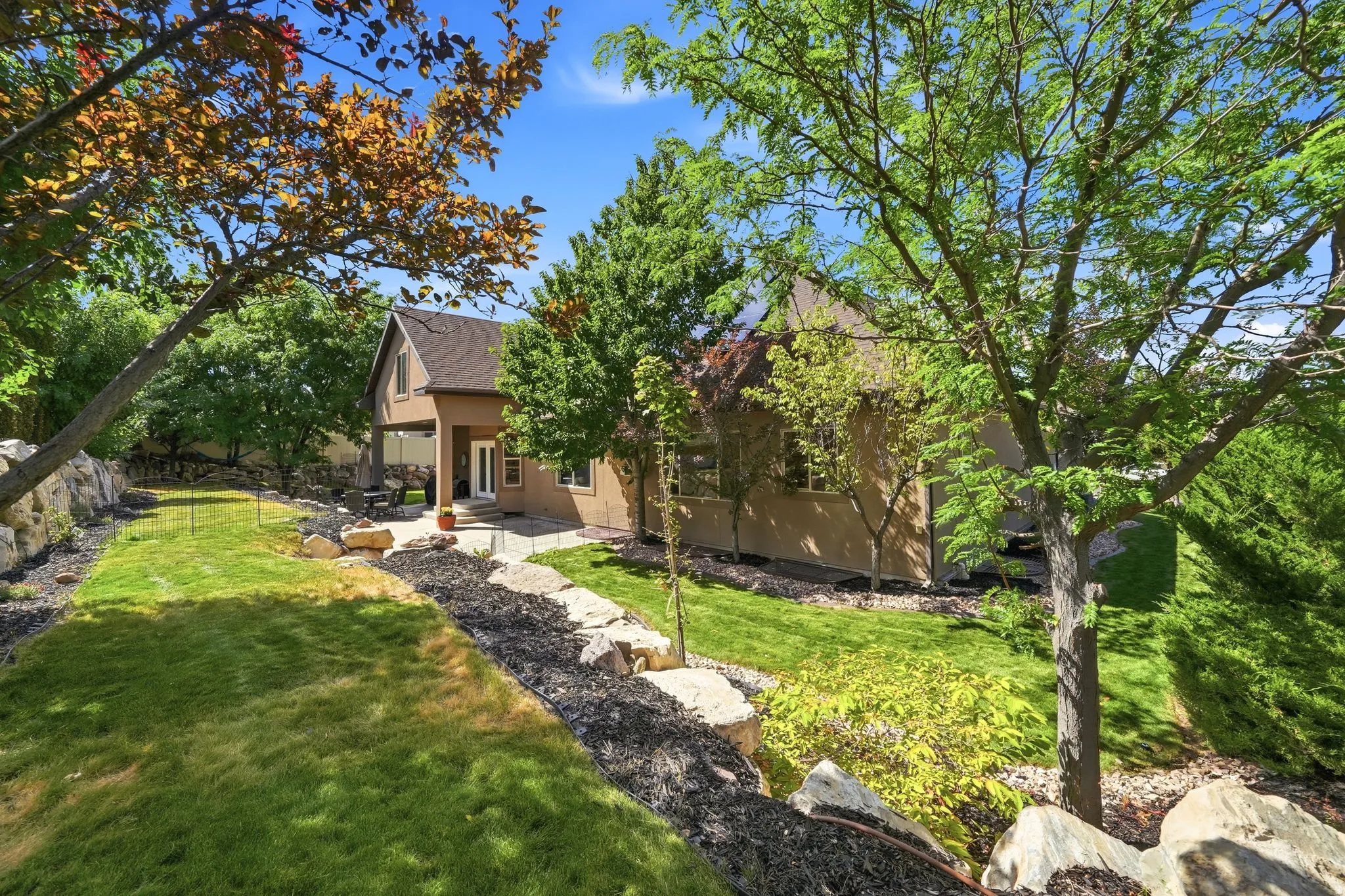Expansive backyard featuring a stone patio, covered outdoor living space, natural rock retaining walls, and mature landscaping