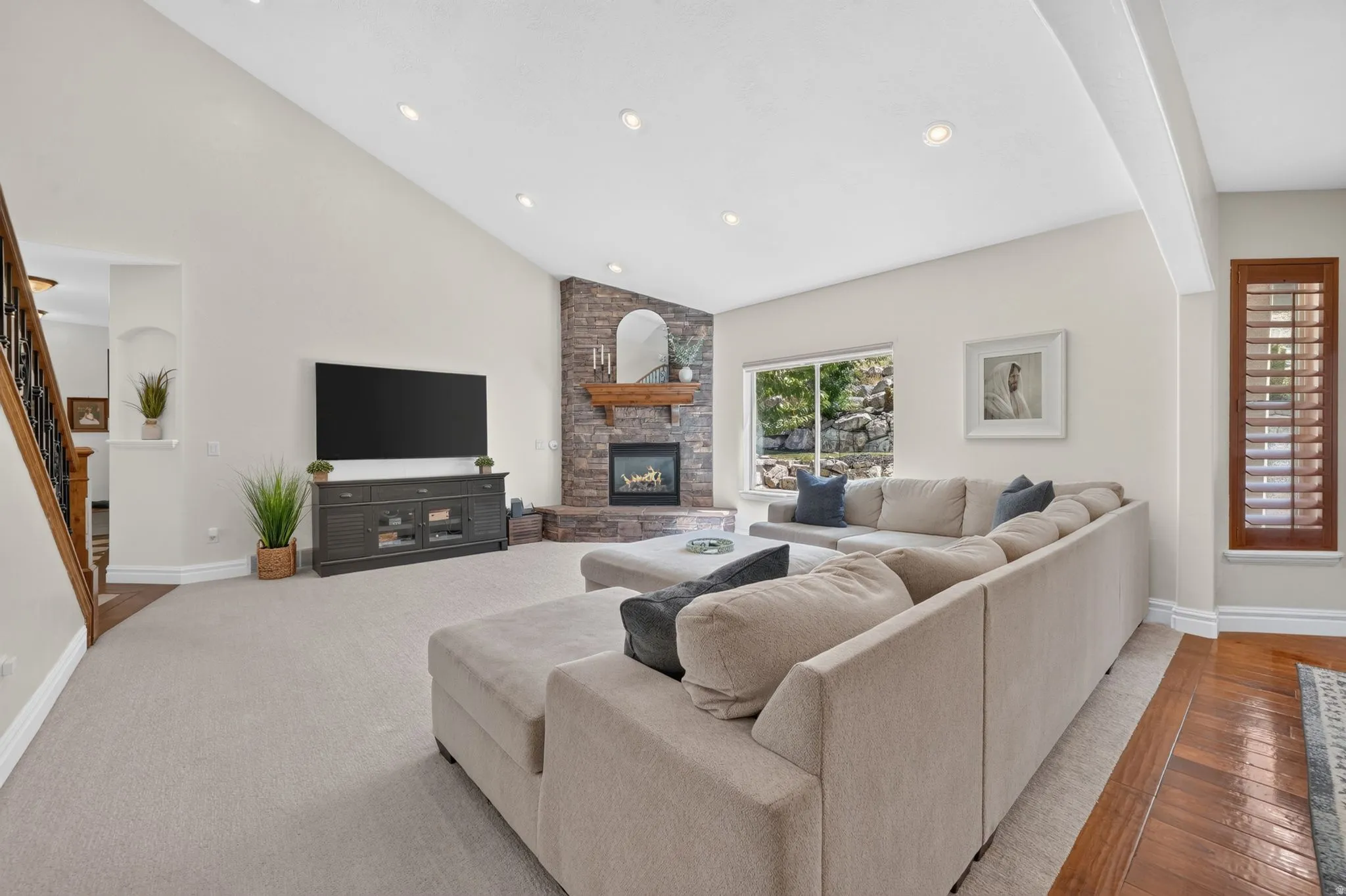 Vaulted ceiling with recessed lighting, stone-clad fireplace with wood mantel, hardwood flooring, and a large window with exterior greenery