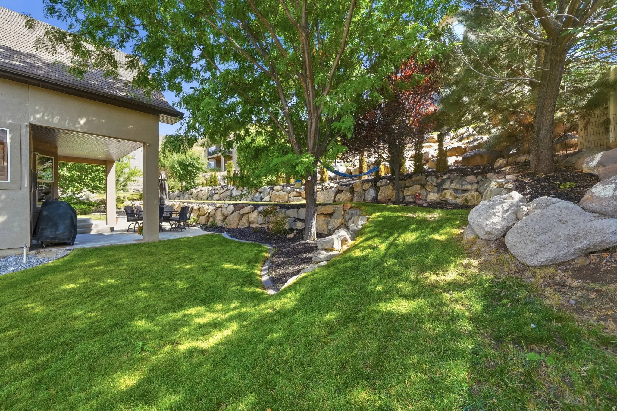 Expansive tiered backyard featuring a manicured lawn, concrete patio, covered outdoor living space, and stone retaining walls